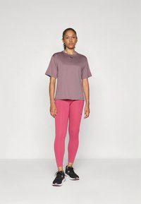 Lilla T-skjorte med korte ermer, rosa leggings, svarte treningssko. Passformen er ledig, og materialene ser lette og pustende ut.