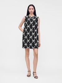 Femme portant une robe noire sans manches avec un motif en treillis de nœuds blancs et des sandales noires à talons hauts, debout devant un fond blanc.