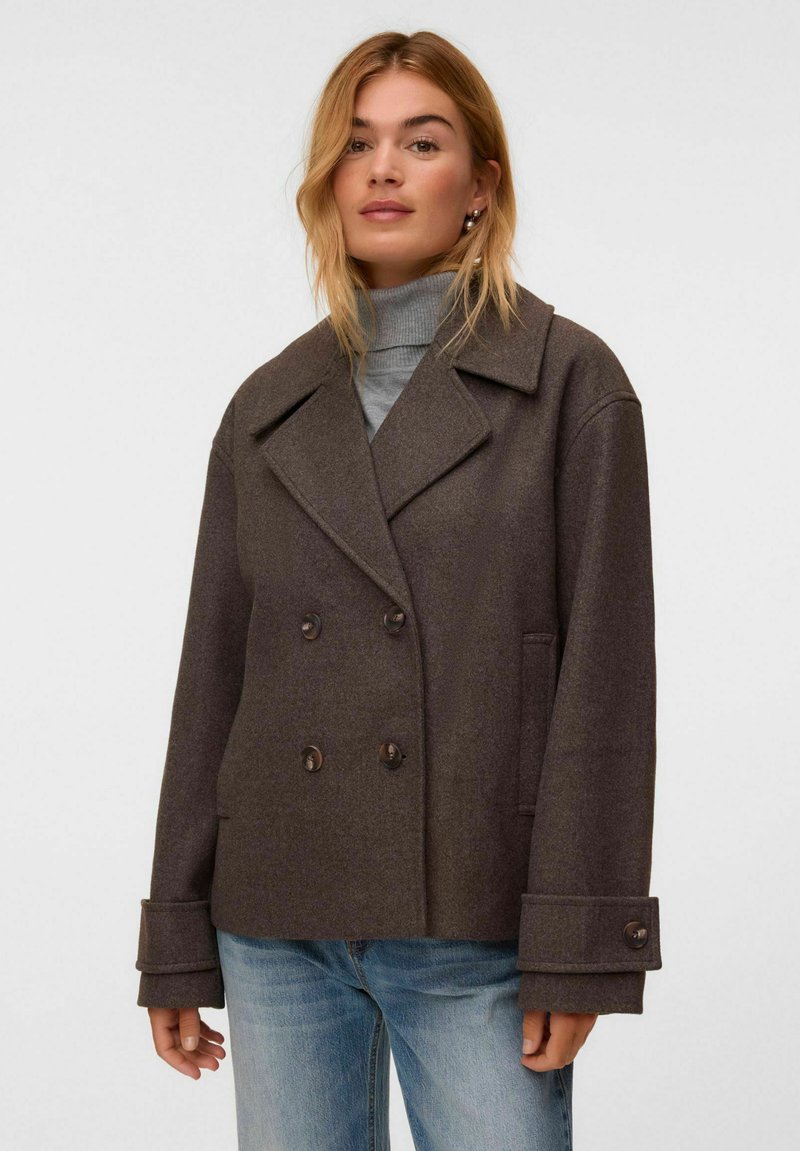 Femme portant un manteau croisé marron foncé sur un pull à col roulé gris et un jean bleu, debout devant un fond clair uni.