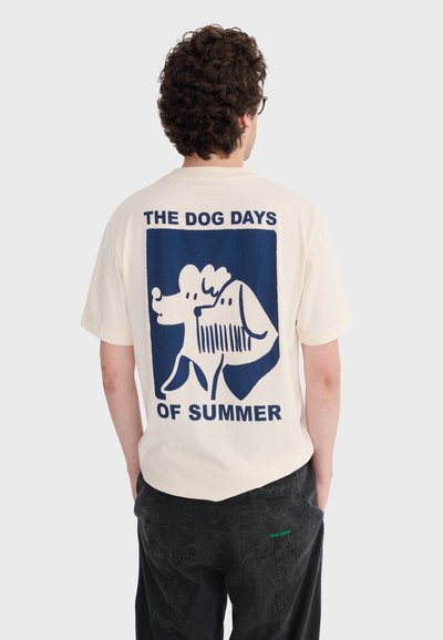 Homme aux cheveux bouclés portant un t-shirt crème avec un graphique bleu de deux chiens et le texte « LES JOURNÉES CANICULAIRES DE L'ÉTÉ » au dos.