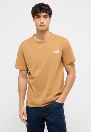 Junger Mann mit kurzen lockigen Haaren, der ein hellbraunes T-Shirt mit Mustang-Logo und dunkelblaue Jeans trägt, steht mit einer Hand in der Tasche.