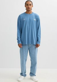 Camiseta de algodón azul de manga larga con estampado gráfico blanco, combinada con jeans azul claro y zapatillas blancas. Estilo casual, ajuste relajado.