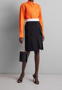 Oranssi pitkähiha, jossa on nappikaulus, yhdistettynä mustiin hameeseen, jossa on valkoinen vyötärökaistale ja sivuhalkio. Musta käsilaukku.