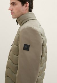Khaki pūkinis švarkas su tekstūruotais smėlio spalvos rankovėmis, aukšta apykakle ir juodu logotipu ant peties. Yra priekinis užtrauktukas ir šoninės kišenės.