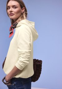 Vrouw draagt een crème hoodie en een sjaal met luipaardprint, met haar hand in de zak en een bruine, getextureerde handtas over de schouder, tegen een lichtblauwe achtergrond.