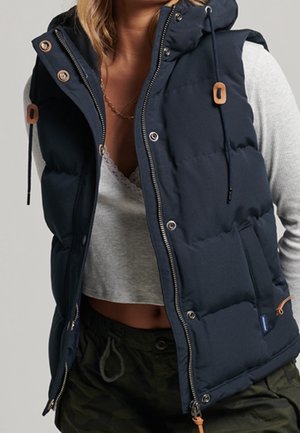 Vrouw draagt een navyblauwe gewatteerde bodywarmer over een lichtgrijs cropped topje en donker groene broek met trekkoord in de taille.