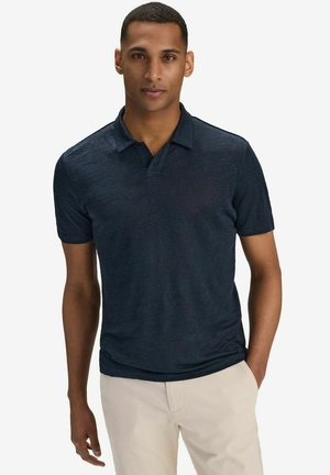 Man som bär en mörkblå pikétröja med kortärmar och ljusbeige byxor, stående med ena handen i fickan mot en vit bakgrund.