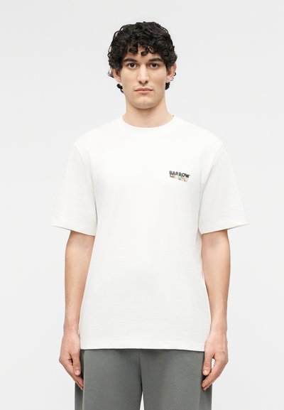 Νεαρός με κατσαρά μαλλιά που φοράει ένα λευκό T-shirt με το λογότυπο Barrow και γκρι παντελόνι, στέκεται μπροστά σε ένα απλό λευκό φόντο.