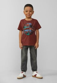 Bordowa bawełniana koszulka z okrągłą grafiką przedstawiającą palmy i scenę zachodu słońca. Zestawiona z szarymi zwężającymi się spodniami i białymi trampkami.