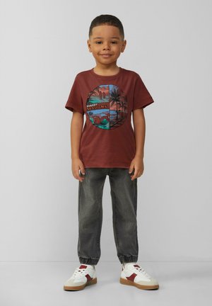 Bordeauxfarbiges Baumwoll-T-Shirt mit einem kreisförmigen Grafikdesign, das Palmen und eine Sonnenuntergangsszene zeigt. Kombiniert mit grauen, schmal zulaufenden Hosen und weißen Sneakern.