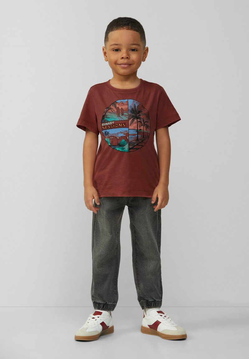 Bordowa bawełniana koszulka z okrągłą grafiką przedstawiającą palmy i scenę zachodu słońca. Zestawiona z szarymi zwężającymi się spodniami i białymi trampkami.
