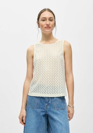 Femme à la peau claire et aux cheveux bruns portant un haut blanc en dentelle sans manches et un jean bleu en denim, debout devant un fond blanc uni.