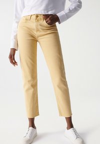 Pantalones de mezclilla amarillos con un corte ajustado, tobillos recortados y bajos deshilachados, combinados con zapatillas blancas y una camiseta de manga larga blanca.