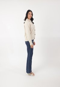 Lichtbeige cardigan met een kraag en houten knopen, gecombineerd met donkerblauwe jeans en hogere sandalen, staande tegen een witte achtergrond.