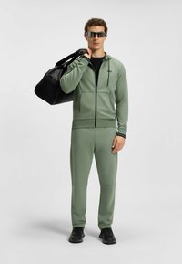 Grüner sportlicher Trainingsanzug aus glattem Material, mit einer Reißverschlusskapuze und schmal zulaufenden Hosen. Schwarze Sporttasche und sportliche schwarze Schuhe.