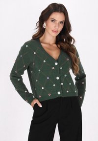 Cardigan tricotat verde cu decolteu în V, având un model în romb și aplicații colorate din strasuri, asortat cu pantaloni negri.