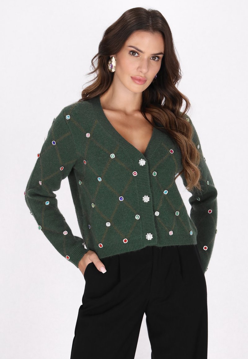Grüne gestrickte Strickjacke mit V-Ausschnitt, verziert mit einem Rautenmuster und bunten Strasssteinen, kombiniert mit schwarzen Hosen.