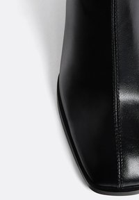 Schwarze Lederstiefelette mit einer markanten, quadratischen Zehenform, glänzender Oberfläche und minimalen Nähten entlang der Naht. Flache Sohle für Stabilität.