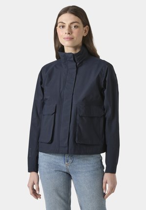 Mujer con chaqueta azul marino de cuello alto con bolsillos delanteros con solapa y vaqueros azul claro, de pie contra un fondo blanco.