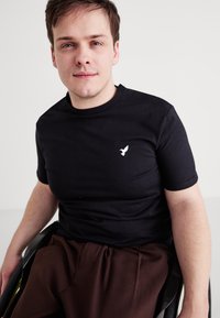 Μαύρο κοντομάνικο μπλουζάκι από βαμβάκι, που διαθέτει ένα μικρό λευκό λογότυπο με πουλί στο αριστερό στήθος. Το μοντέλο κάθεται σε αναπηρικό καροτσάκι.