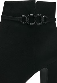Bottine en daim noir avec une fine lanière et quatre anneaux métalliques circulaires liés au-dessus du talon.