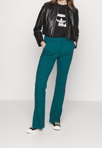 Musta nahkainen cropped-takki, istuva tumma t-paita, jossa on graafinen printti, sekä teal-väriset leveälahkeiset housut, yhdistettyinä mustiin ja valkoisiin lenkkareihin.