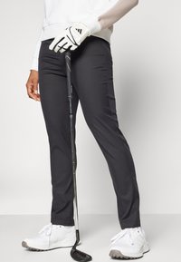 Μαύρο, ραμμένο παντελόνι γκολφ συνδυασμένο με άσπρο μακρυμάνικο τοπ και λευκά αθλητικά παπούτσια. Μια ράβδος γκολφ κρατιέται σε ένα χέρι, με λευκό γάντι.