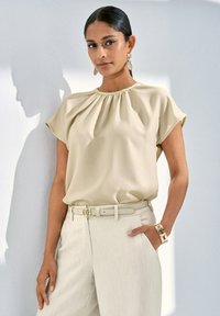 Beige, kurzärmliges Oberteil mit gerafftem Ausschnitt, kombiniert mit hellen, weiten Hosen und einem schmalen Gürtel. Auffälliger Schmuckakzent.