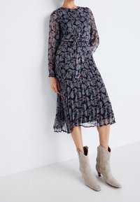 Blommönstrad klänning med lång ärm i mörka nyanser, med en lager-på-lager, veckad kjoldetalj. Matchas med spetsiga beige mocka ankelstövlar.