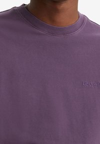 Osoba v fialovém tričku s kulatým výstřihem značky Levi's s vyšitým logem na levé hrudi, zobrazená od krku do poloviny trupu.