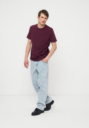 Muž se mírně naklání vpřed s jednou rukou v kapse, nosí tmavě fialové tričko, světlé modré džíny a černé boty, stojí na bílé podlaze.