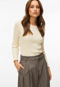 Femme aux longs cheveux bruns portant un pull en maille torsadée crème et un pantalon plissé gris taille haute avec ceinture, posant avec une main dans la poche.