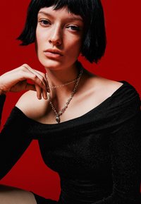 Czarny top typu off-shoulder z połyskiem, srebrny naszyjnik z łańcuszkiem z zawieszką w kształcie czaszki, modelka pozując na czerwonym tle.