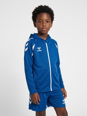 Dreng med krøllet hår iført en blå hummel sportshættetrøje og matchende shorts, der står mod en ensfarvet baggrund.