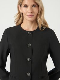 Woman wearing a black blazer with four decorative floral buttons and small silver hoop earrings against a plain light background.