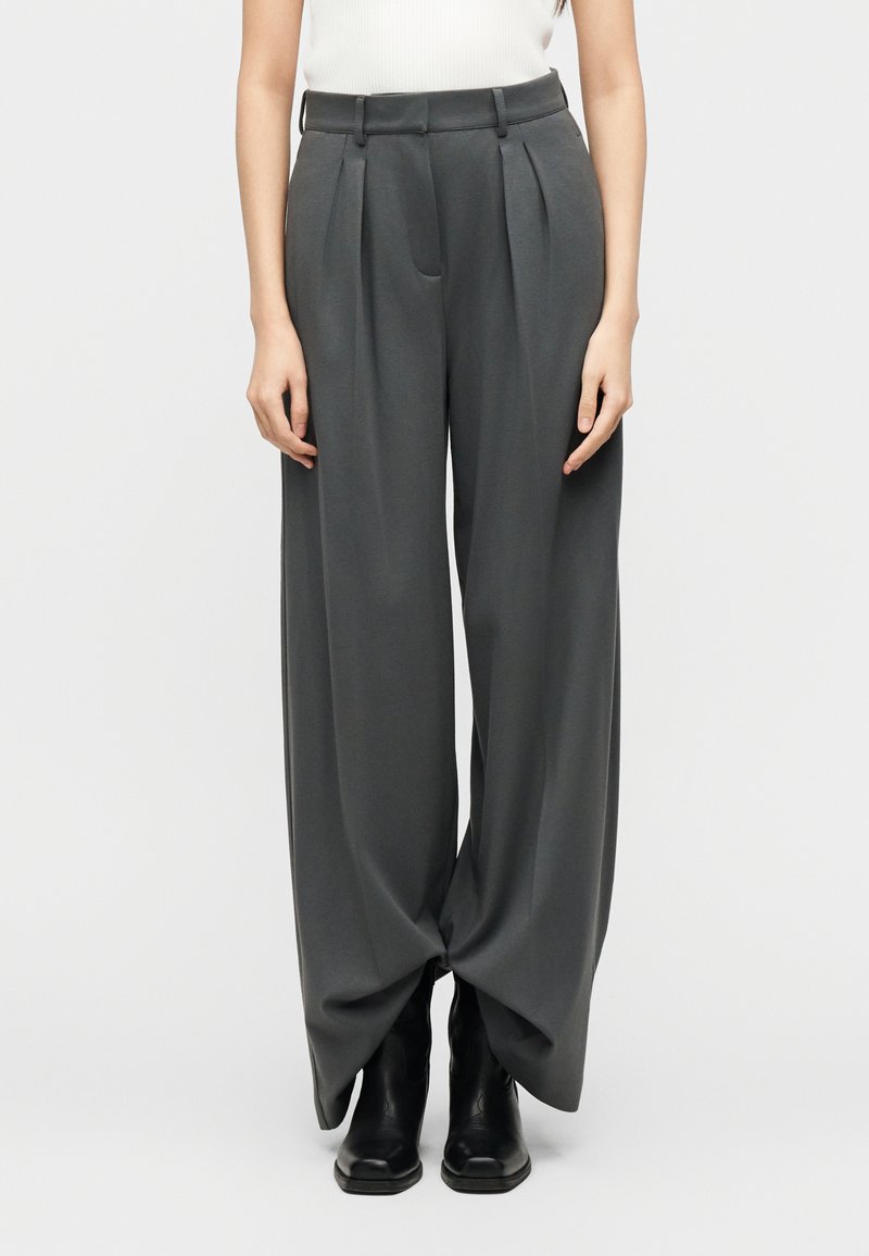 Γυναίκα που φοράει ψηλόμεσο, φαρδύ σκούρο γκρι π αντελόνι με πιέτες, σε συνδυασμό με μαύρες μπότες και λευκό αμάνικο τοπ, στέκεται μπροστά σε λευκό φόντο.