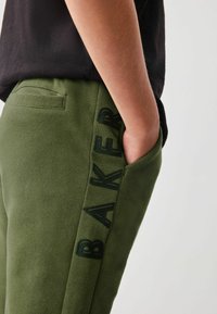 Zelena trenirka z gladko teksturo, stransko žepom in vertikalnim logotipom "BAKER" v temnejši zeleni barvi na levi nogi.