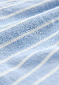 Lichtblauwe stof met witte verticale strepen, met een gestructureerde weving. Het oppervlak vertoont subtiele variaties in kleur en patroon.