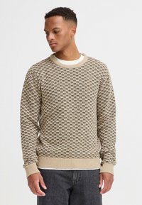 Suéter de punto beige con una textura estampada y puños acanalados. Cuello redondo y mangas largas, llevado sobre una camisa blanca.