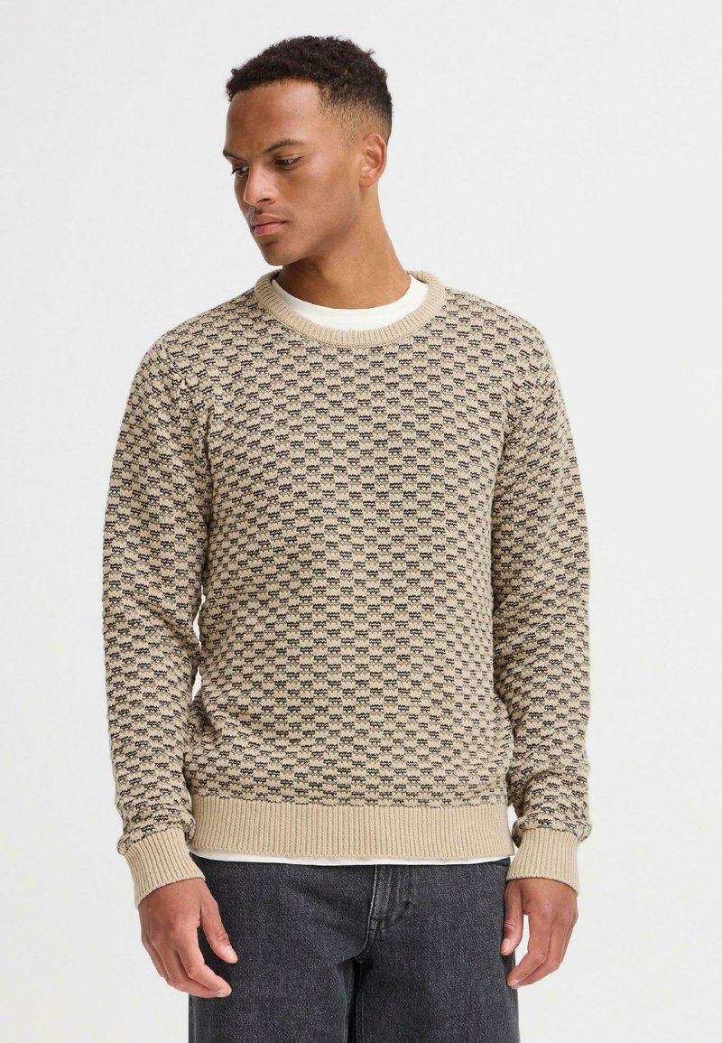 Suéter de punto beige con una textura estampada y puños acanalados. Cuello redondo y mangas largas, llevado sobre una camisa blanca.