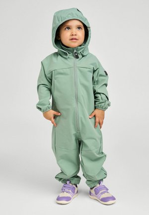 Kleinkind trägt einen hellgrünen, wasserdichten Overall mit Kapuze und lila Turnschuhe, steht und blickt leicht nach oben.