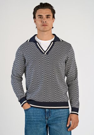 Maglione a motivo chevron in navy e crema con scollo a V e colletto a contrasto. Polsini e orlo a coste, indossato sopra una t-shirt bianca, abbinato a jeans blu.