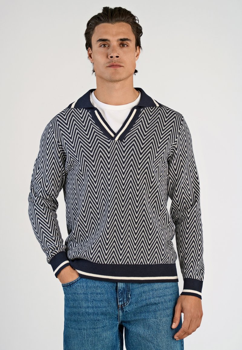 Maglione a motivo chevron in navy e crema con scollo a V e colletto a contrasto. Polsini e orlo a coste, indossato sopra una t-shirt bianca, abbinato a jeans blu.