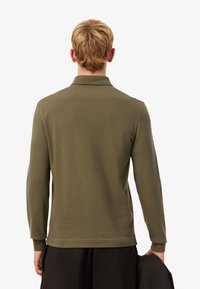 Olivgrön långärmad pikétröja med krage, med en mjuk textur och figurnära design. Bakifrån ses en rak nederkant och långa ärmar.