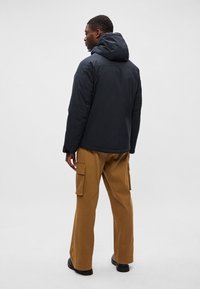 Jaqueta preta com capuz, fechamento com zíper, textura suave e corte descontraído, combinada com calças cargo castanho-claras e sapatos escuros.