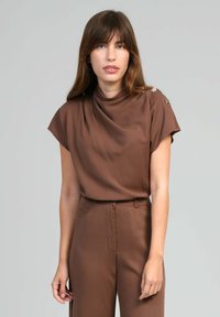 Femme avec des cheveux bruns raides portant un haut marron à manches courtes avec des boutons sur les épaules et un pantalon taille haute assorti, sur fond gris.