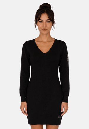Donna con capelli scuri raccolti in uno chignon, che indossa un vestito nero aderente a maniche lunghe con scollo a V, in piedi davanti a uno sfondo chiaro e uniforme.