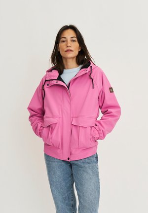 Femme portant une veste à capuche rose avec des poches avant et un jean bleu, debout devant un fond clair uni.