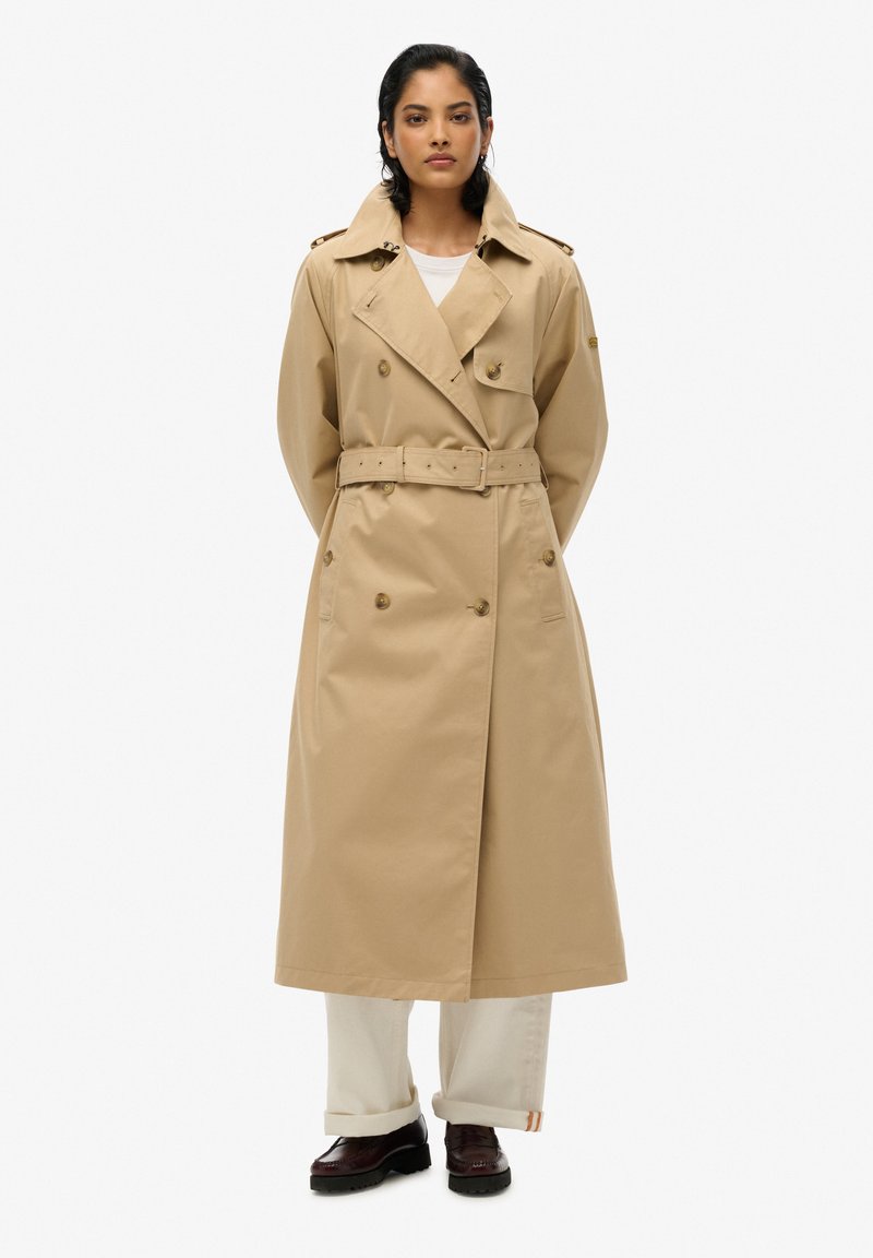 Beżowy trencz z paskiem w talii, podwójnym zapięciem, szerokimi klapami i epauletami na ramionach, wykonany z gładkiego, teksturowanego materiału.