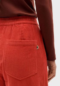 Pantaloni di velluto a coste rossi con superficie testurizzata, dotati di una tasca posteriore e un piccolo dettaglio logo rotondo. La vita è elasticizzata per maggiore comfort.